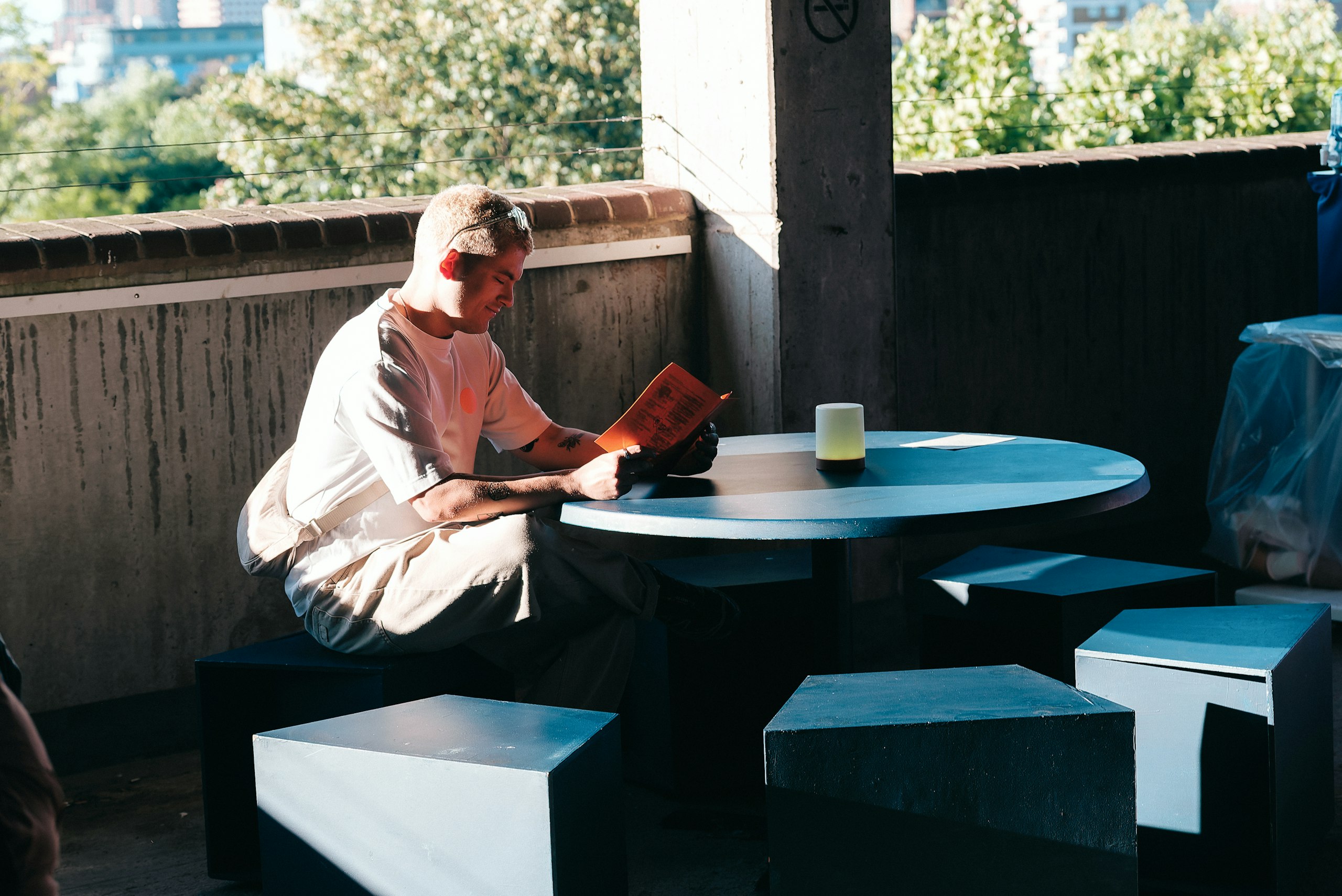 A man in a white T-shirt reads an orange programme at a round table on a sunlit concrete rooftop terrace.