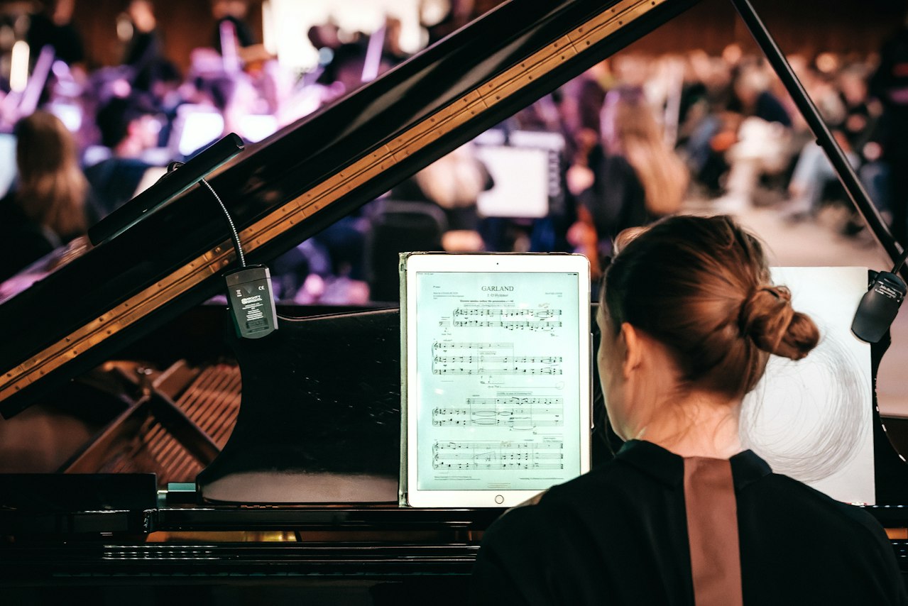 A conductor gestures to an orchestra, seen through the raised lid of a grand piano.