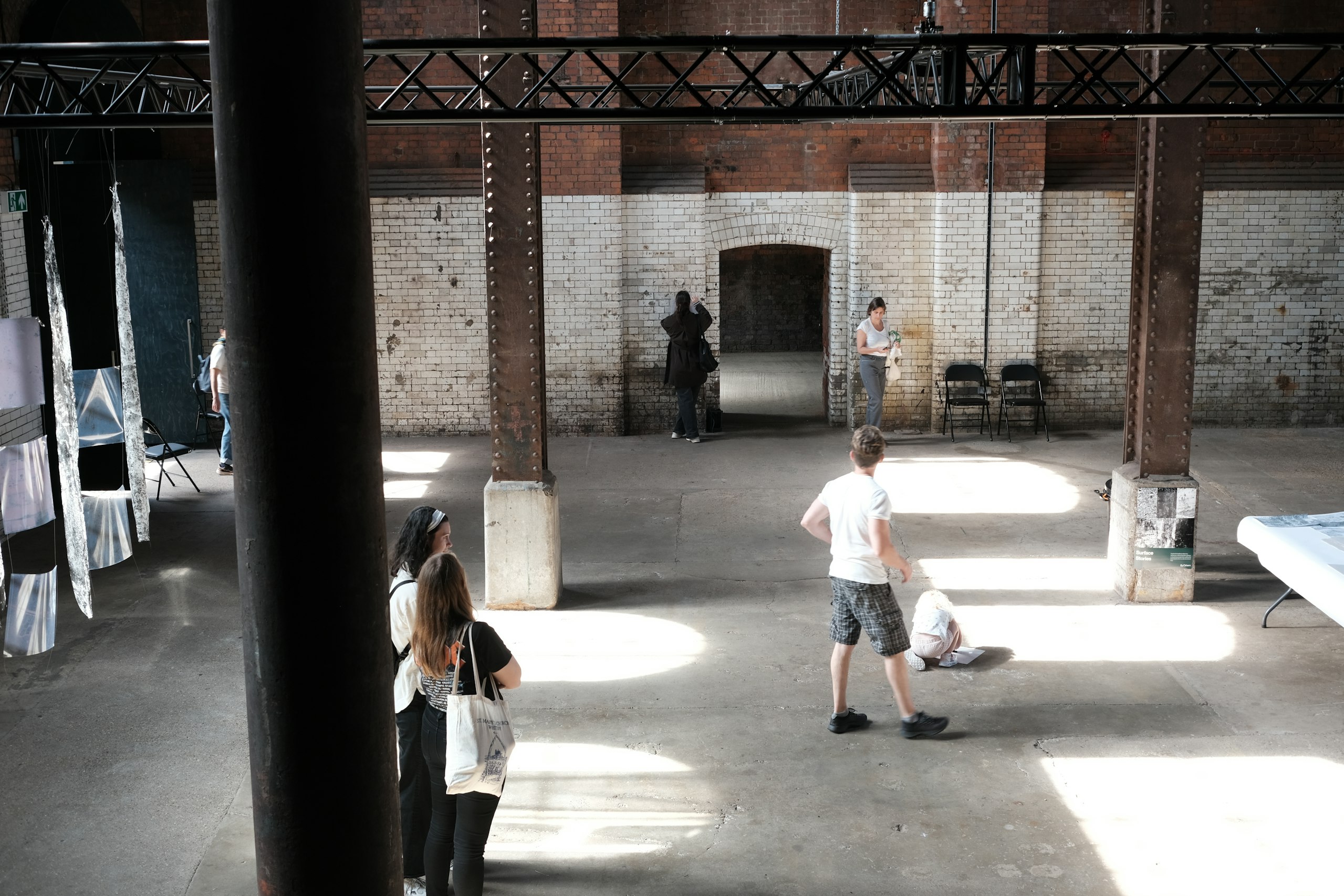 Open house: Visitors explore a sunlit industrial hall with whitewashed brick walls, iron columns and hanging artworks.