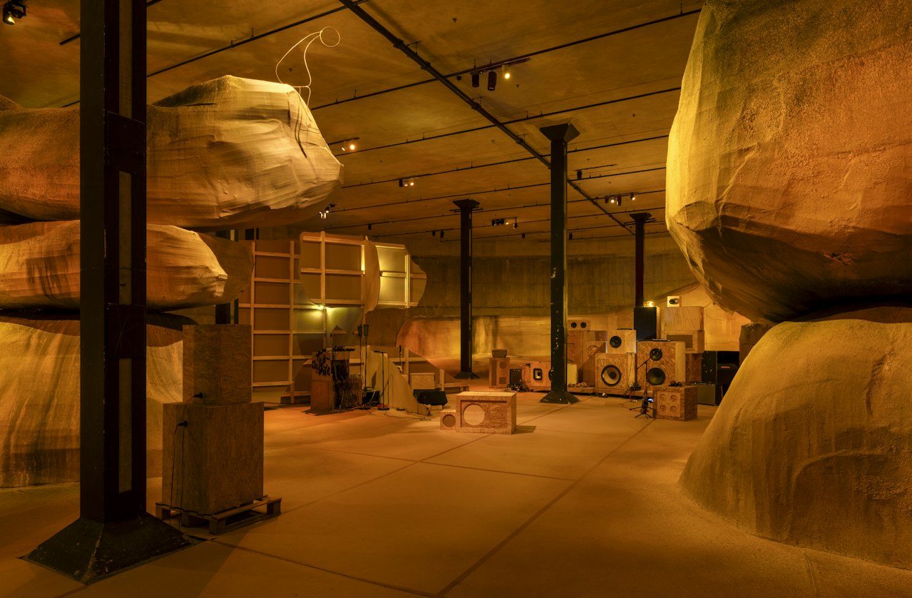 An installation view showing large, rock-like forms constructed from cardboard and timber, with speakers arranged toward the end of the room. The space is illuminated in warm orange light
