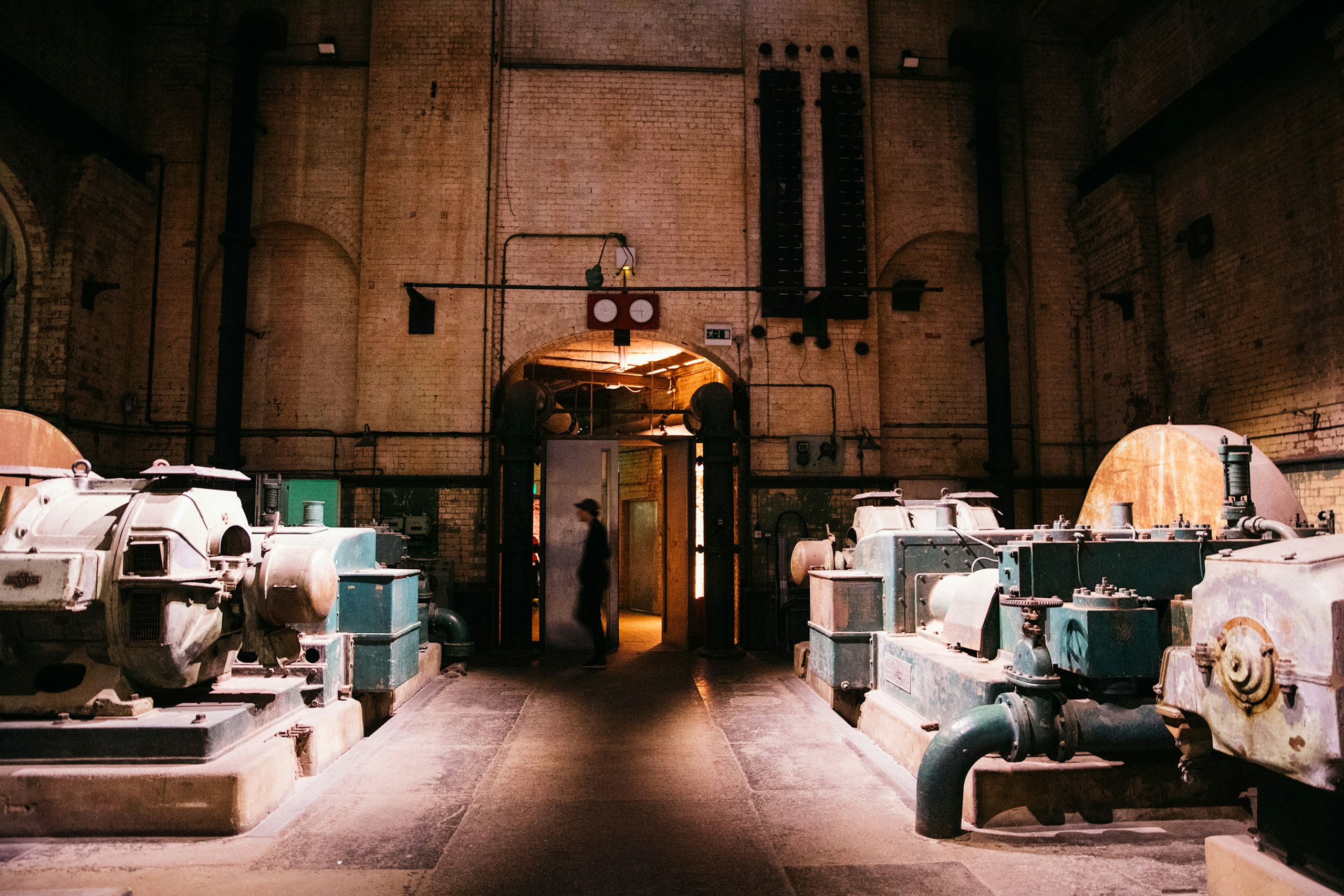 Interior image: A figure walks through a lit doorway at the end of a dim brick hall lined with old teal pumping machinery.