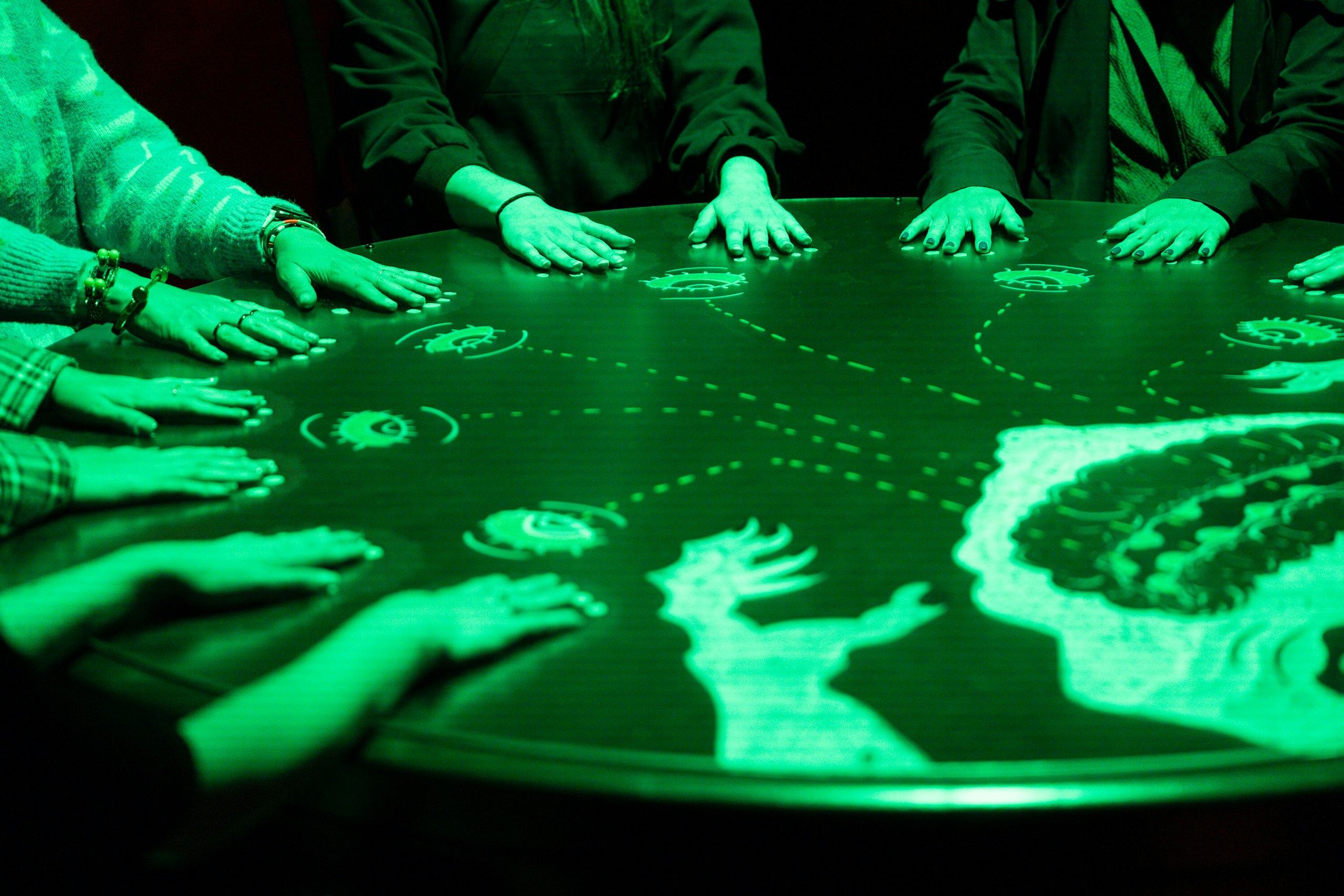 Many hands rest on a glowing green round table projected with illustrated symbols and figures.