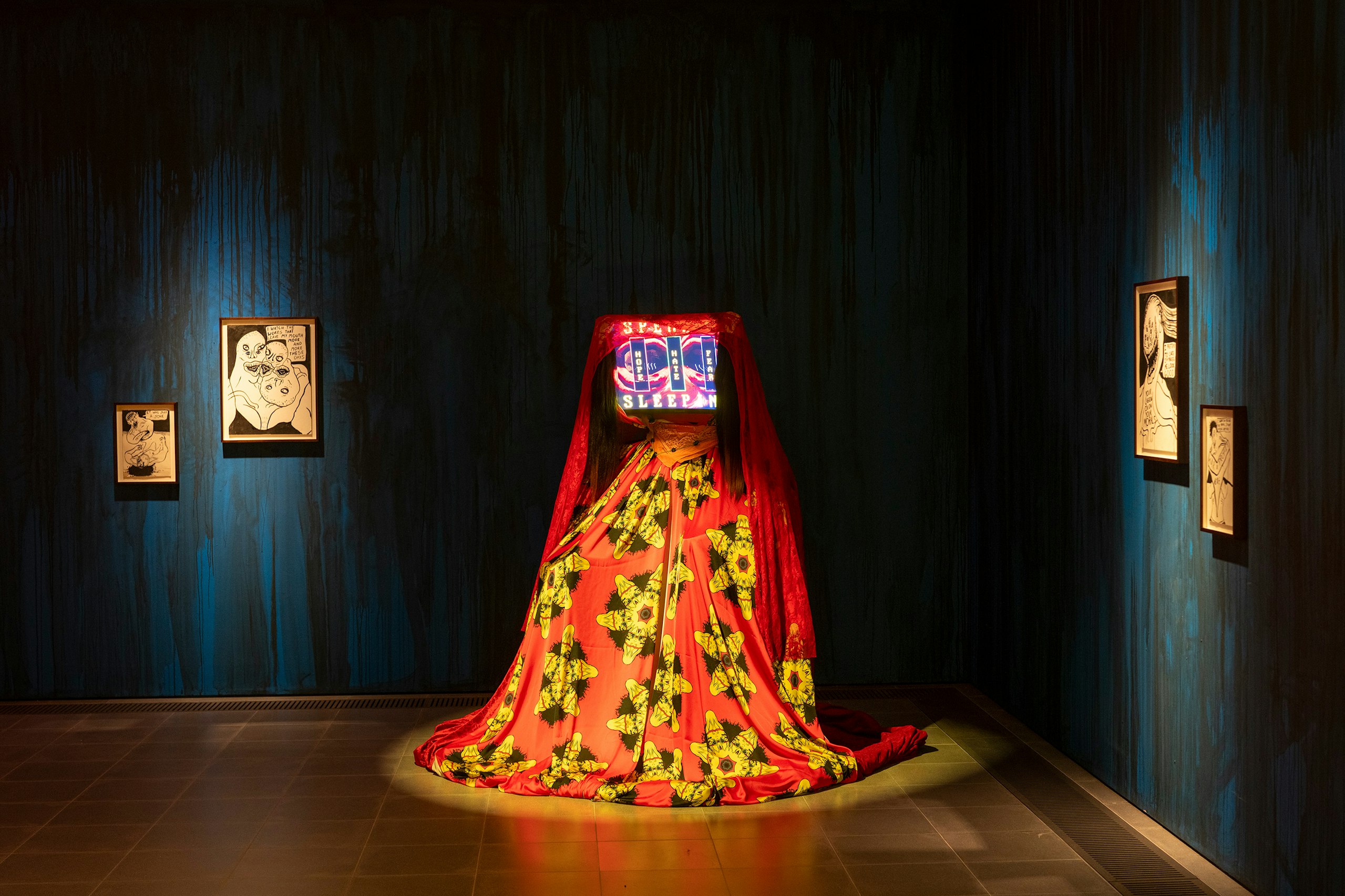 THE DELUSION, Danielle Brathwaite-Shirley:Gallery installation: a veiled figure in a red and yellow patterned gown with a glowing screen for a head, spotlit in a dark blue room hung with framed black-and-white drawings.
