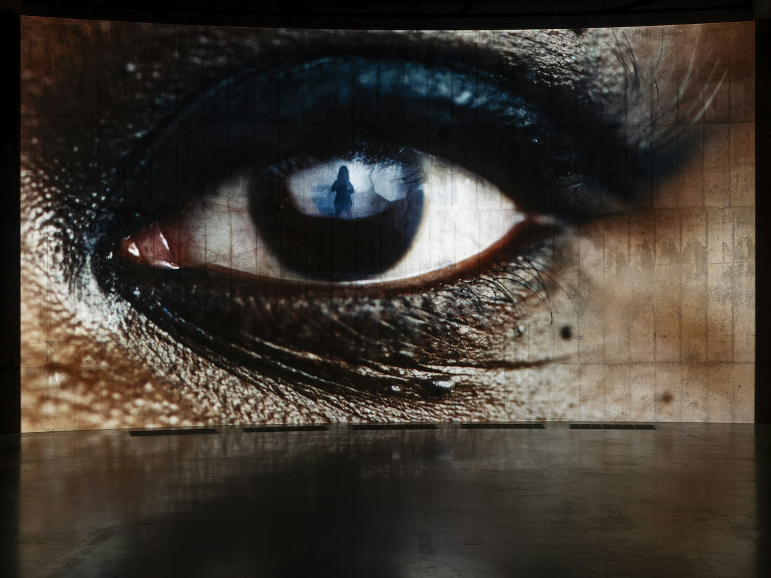 A large projected close-up of an eye fills a curved concrete wall, a small figure reflected in its iris.