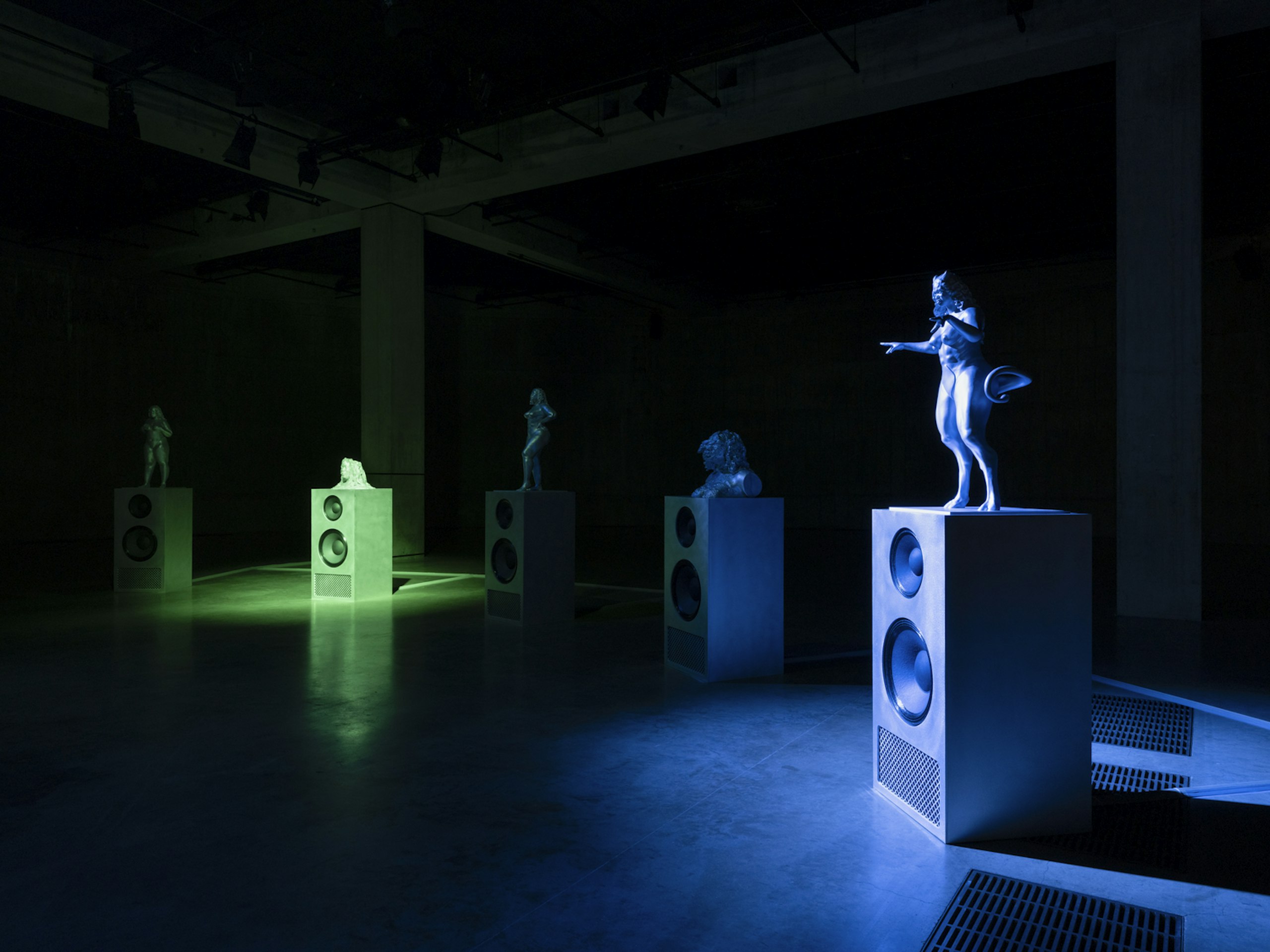 Christelle Oyiri: In a dark gallery, pale figurative sculptures stand on top of loudspeaker cabinets, lit in blue and green.