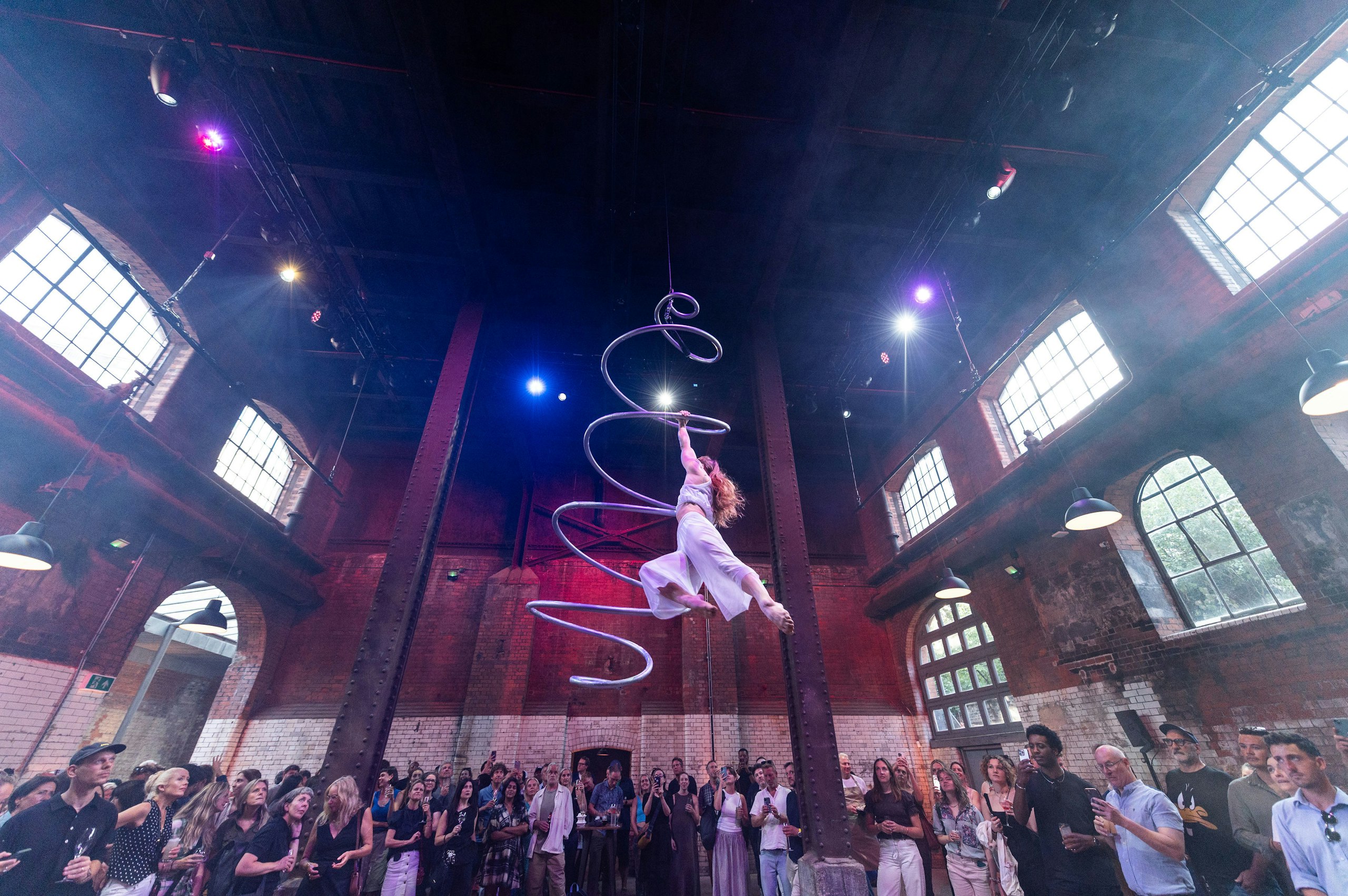An aerial performer hangs from a suspended spiral sculpture in a brick hall with tall arched windows, watched by a crowd below.