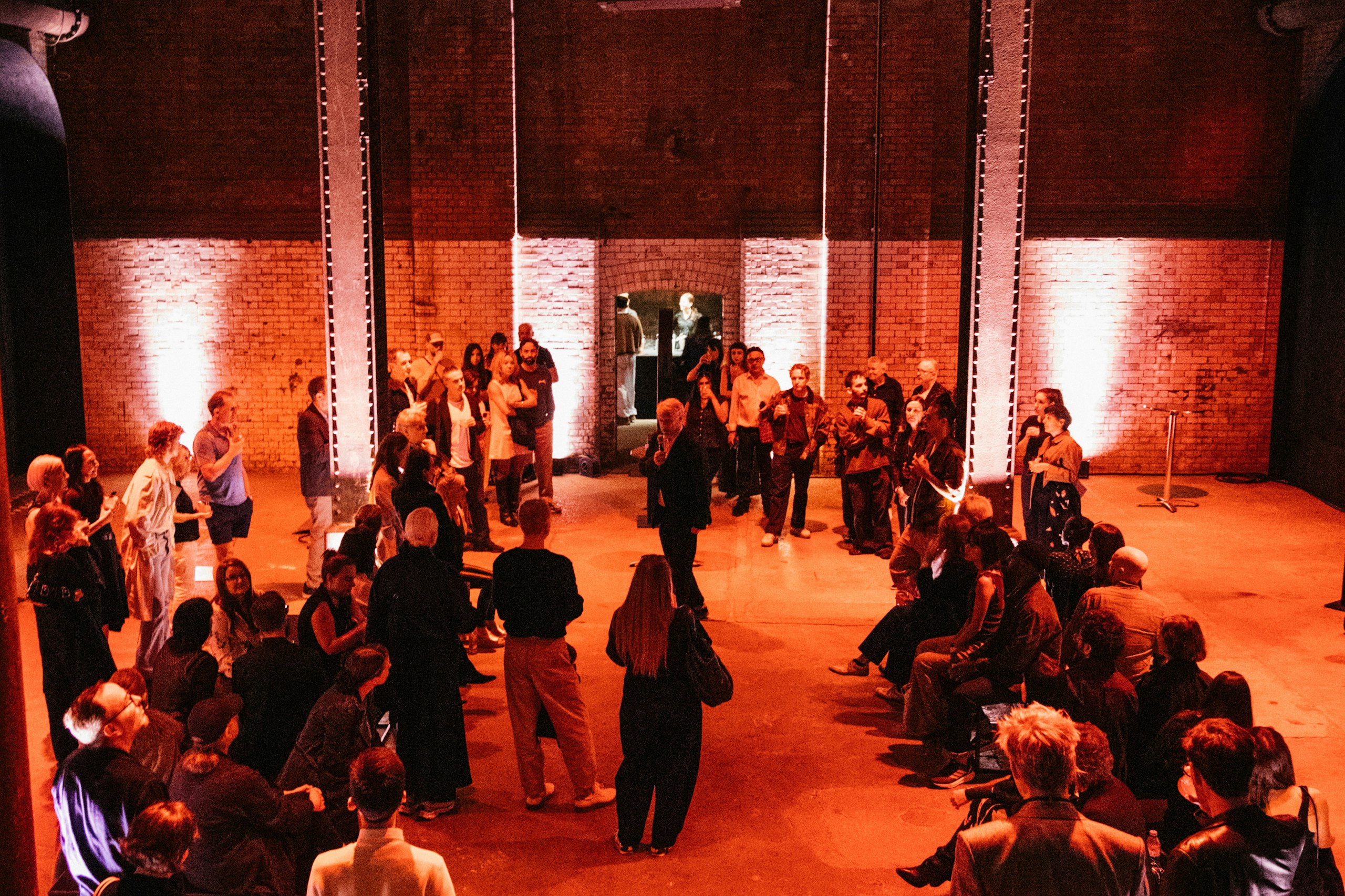 Guests gather at an evening event in an industrial hall lit in warm orange-red, beneath an overhead lighting rig.