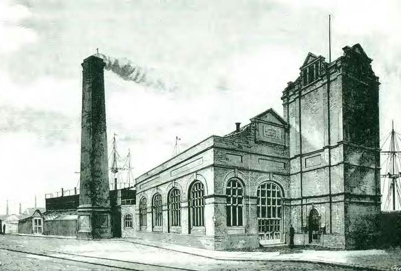 A 19th-century engraving of Wapping Hydraulic Power Station: a tall smoking chimney, an arched-window engine house and a tower, with ships' masts behind.