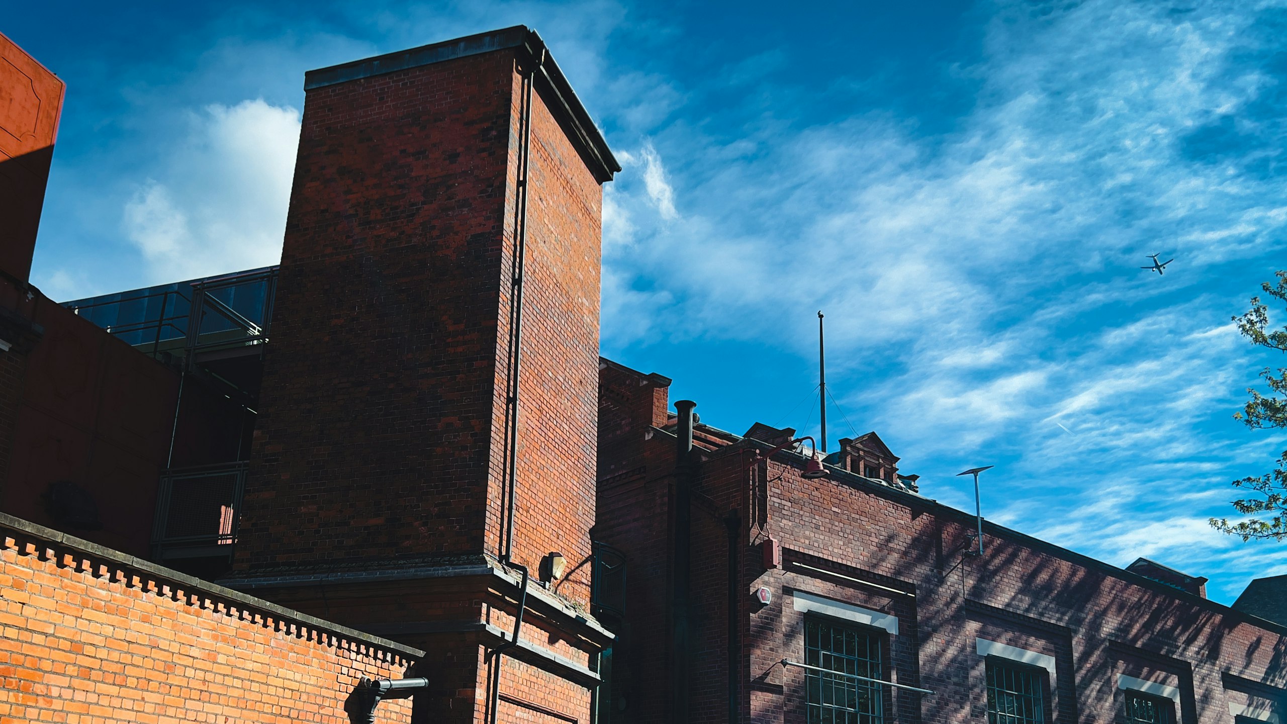 Exterior image: Red-brick industrial buildings and a tall brick tower beneath a bright blue sky, with a plane passing overhead.