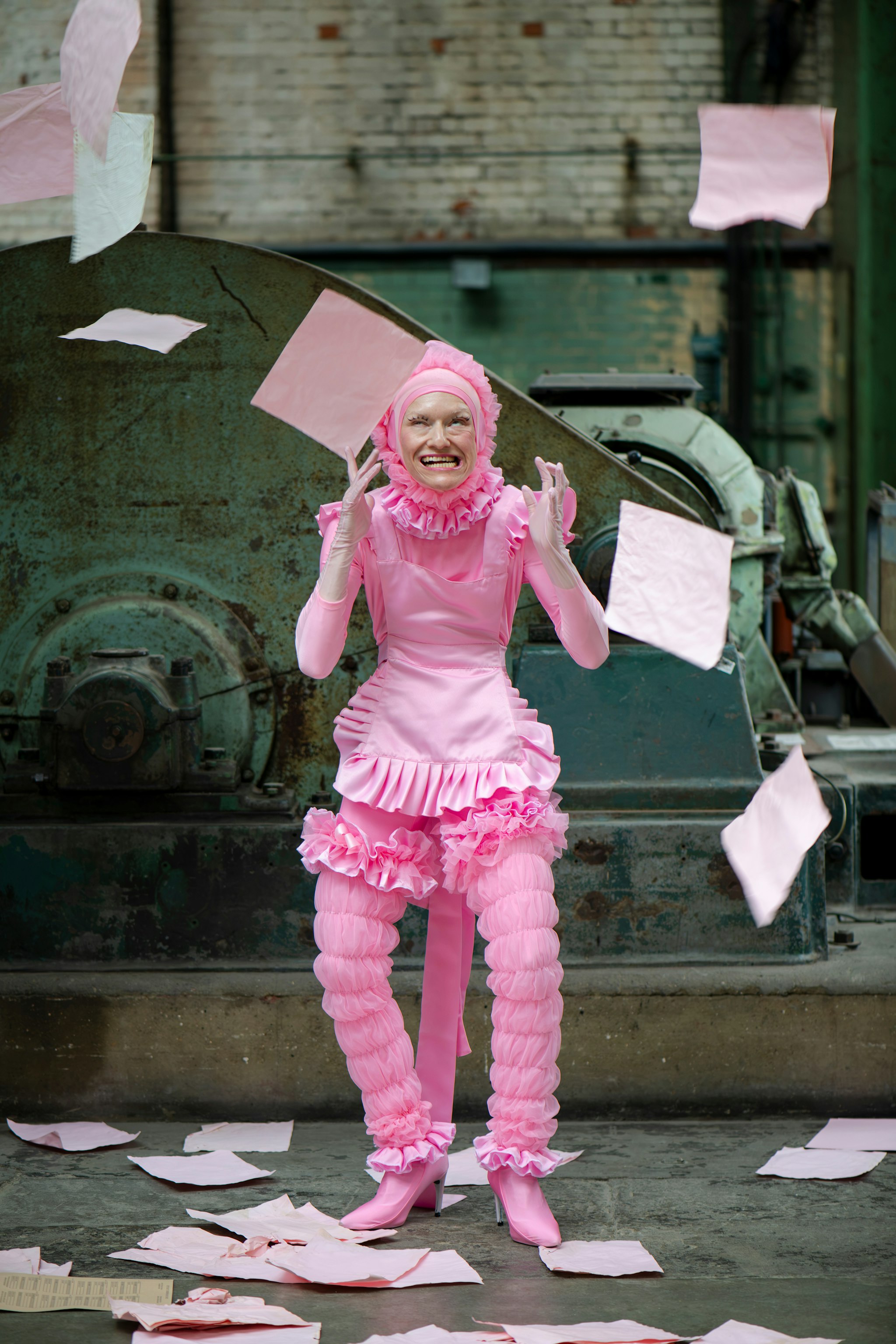 A performer in an elaborate head-to-toe pink costume stands in an industrial space, grimacing with teeth bared and hands raised beside their face. The outfit includes a ruffled hood framing the face, a frilled bib and short skirt, puffy ruched leggings, and pink boots. Sheets of pink paper fly through the air around them and litter the concrete floor. Behind them are a weathered brick wall and large rusted green machinery.